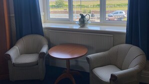 A room with two patterned armchairs, a small wooden table, and a view of a coastal landscape through a window with blue curtains. - The Clarkes Hotel (Barrow-in-Furness)