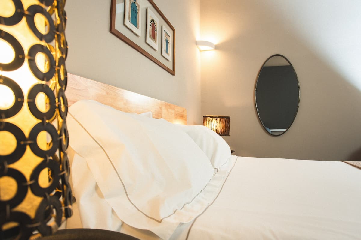 Un lit bien fait, avec du linge de lit blanc, une lampe de chevet et un miroir fixé au mur.