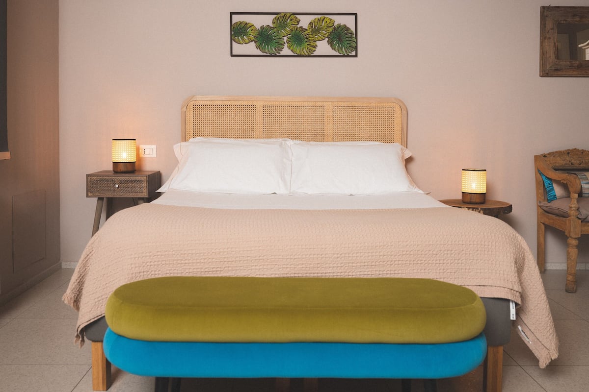 Un lit bien fait, recouvert d’une couverture beige, avec deux lampes de chevet, une tête de lit en bois et un banc coloré placé au pied du lit.