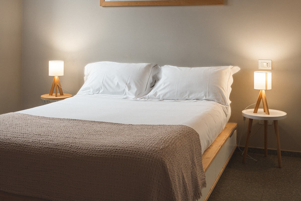 Un lit bien fait, avec du linge de lit blanc et une couverture marron, flanqué de deux tables de chevet sur lesquelles sont posées des lampes.