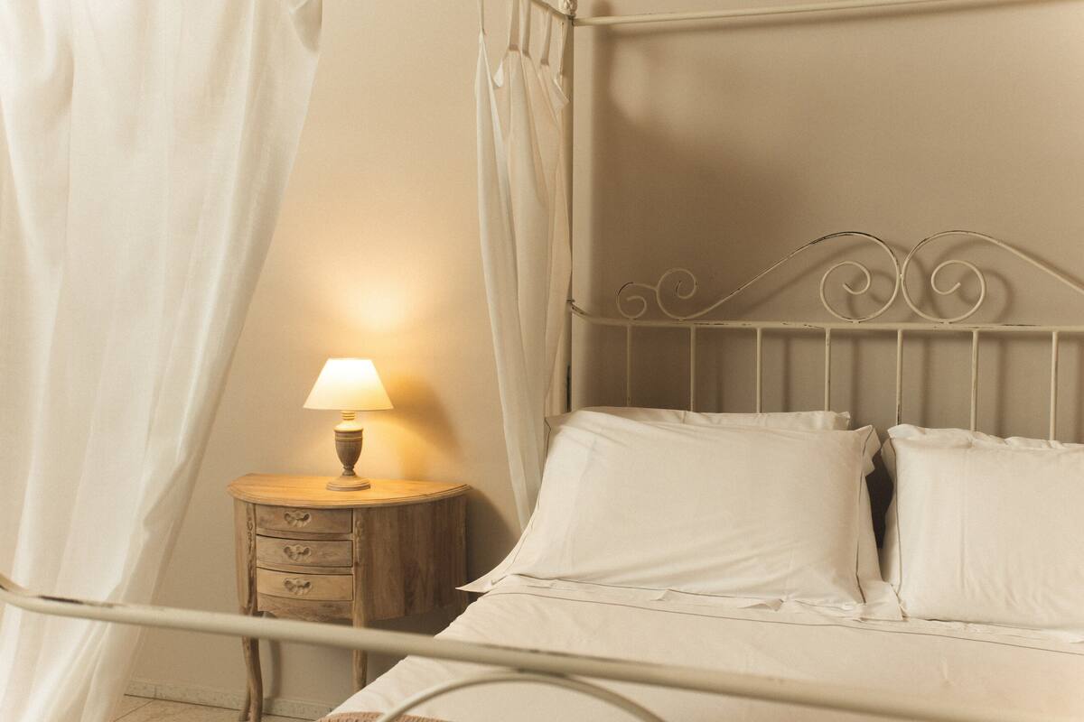 Un lit avec un cadre en métal, une table de chevet avec une lampe et un rideau.