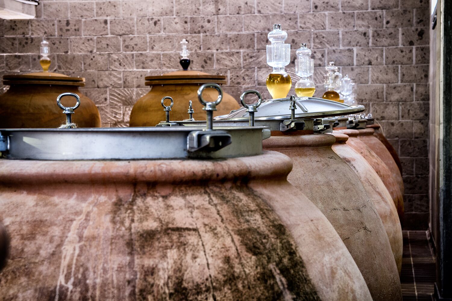 A row of large, rustic wine barrels with metal lids and handles, set against a brick wall background.