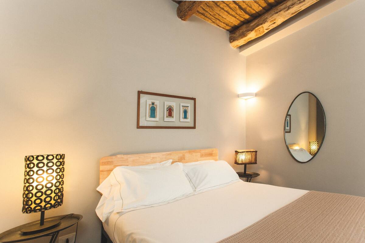 Un lit bien fait, avec du linge de lit blanc, une tête de lit en bois et un plaid beige. Il y a deux lampes de chevet, l’une avec un abat-jour à motifs et l’autre avec un design simple. Un tableau encadré est accroché au-dessus du lit, et un miroir rond est fixé au mur, à droite.
