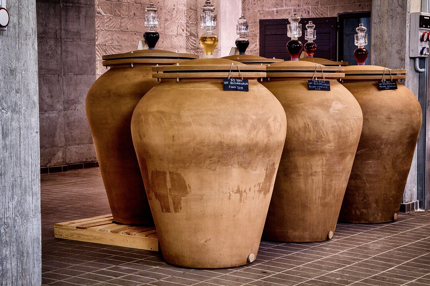 Grote terracotta plantenbakken met labels, een betegelde vloer en een bakstenen muur op de achtergrond.
