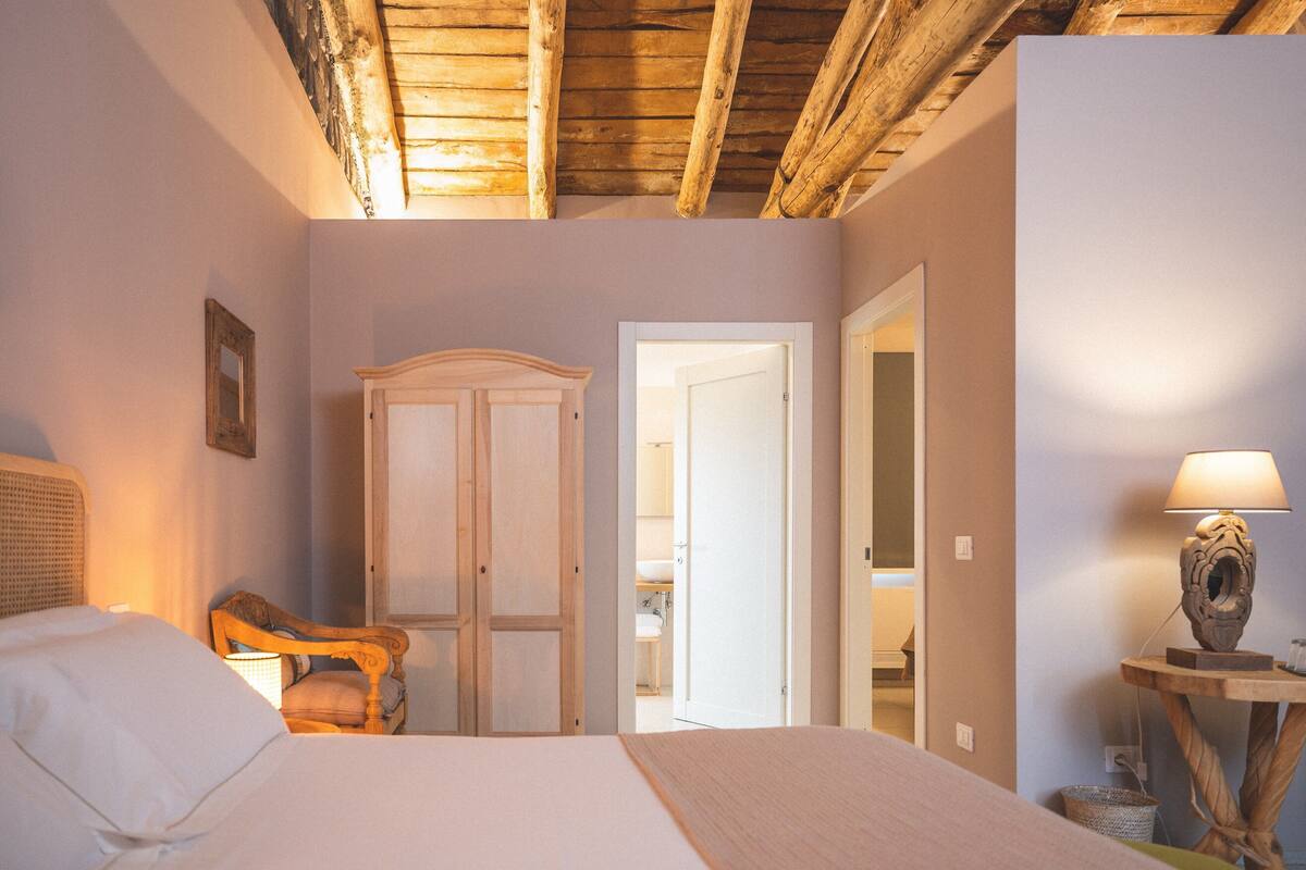 Une chambre à coucher comprenant un lit, une armoire en bois, une chaise, une lampe et une porte donnant sur une autre pièce.