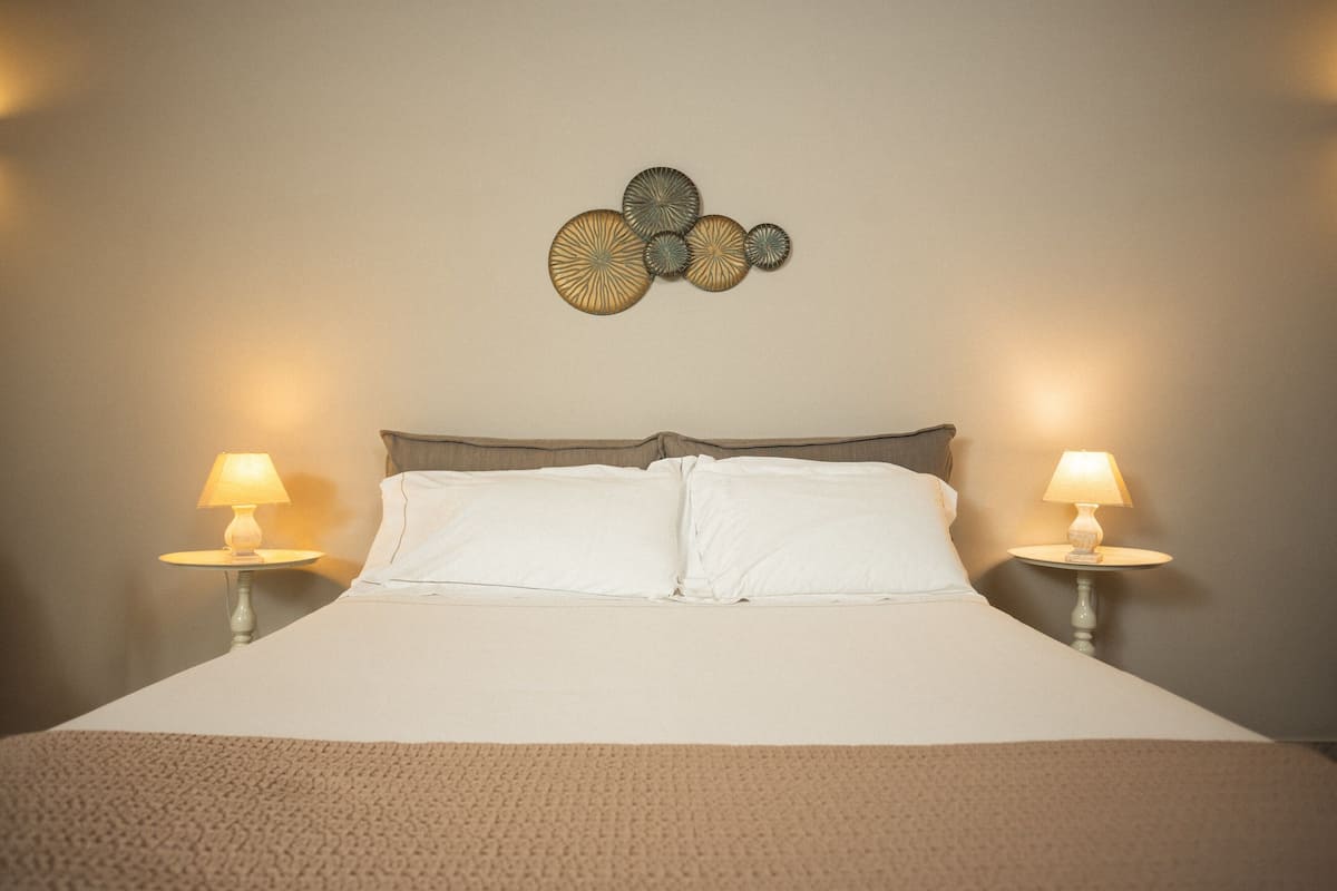 Un lit bien fait, avec deux lampes de chevet, une œuvre d’art accrochée au mur au-dessus, et un plaid à texture intéressante.