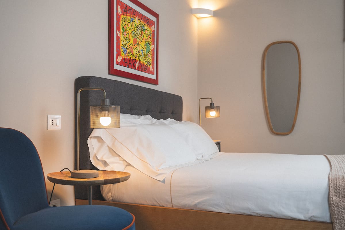 Une chambre d’hôtel avec un lit, une chaise, une petite table, un miroir fixé au mur et une œuvre d’art colorée au-dessus du lit.