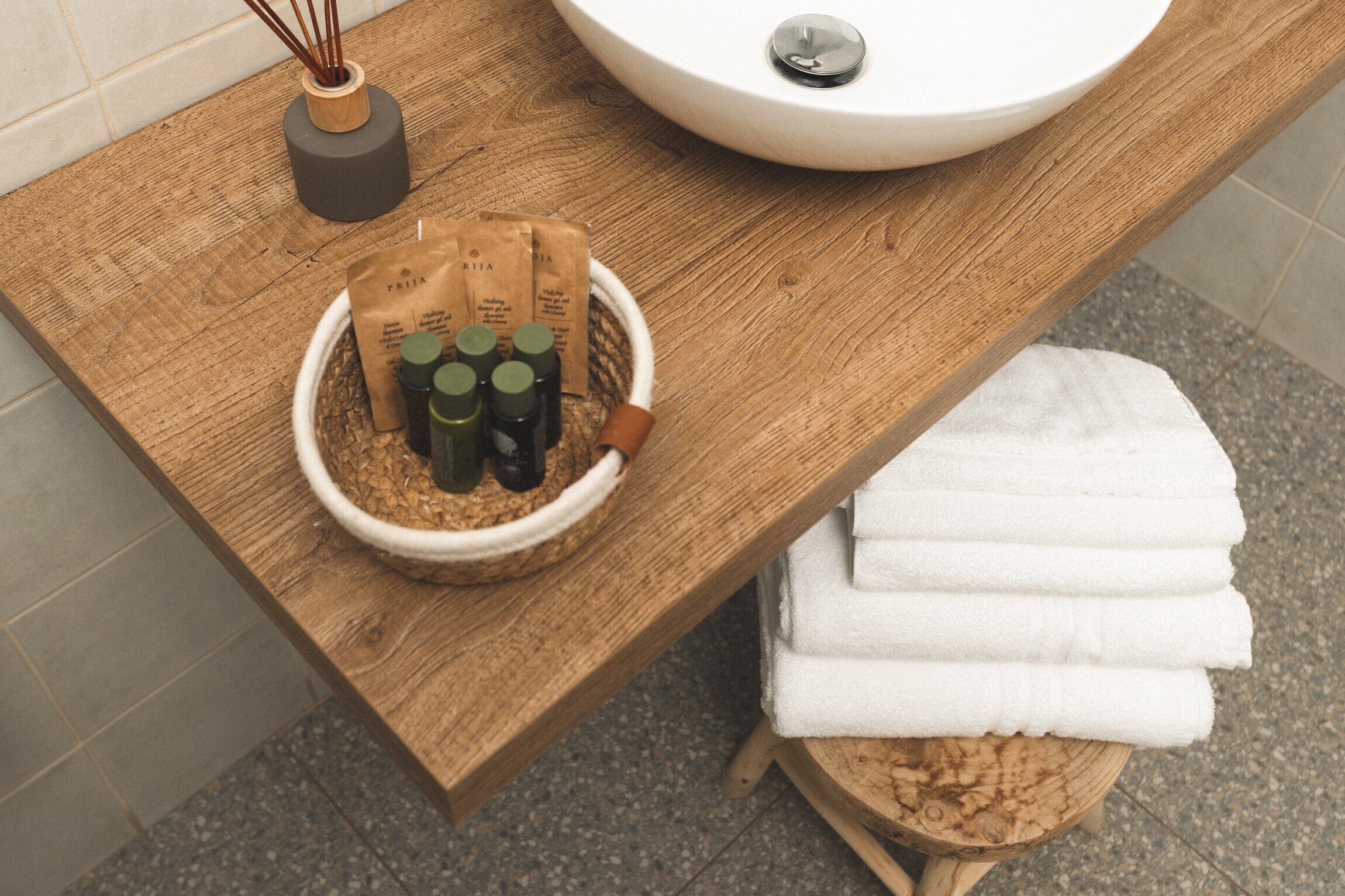 A wooden vanity with a white sink, a bowl of toiletries, and a stack of white towels.