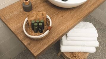 A wooden vanity with a white sink, a bowl of toiletries, and a stack of white towels.