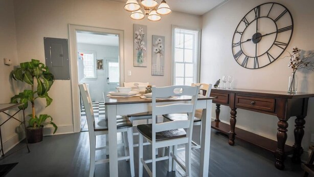 Een eethoek met een witte tafel en stoelen, een houten buffetkast, een grote wandklok en een potplant.