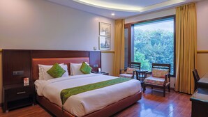 A hotel room with a large bed, a wooden headboard, a nightstand, a desk, a chair, and a window with curtains. - Amber Inn by Orion Hotels (New Delhi)