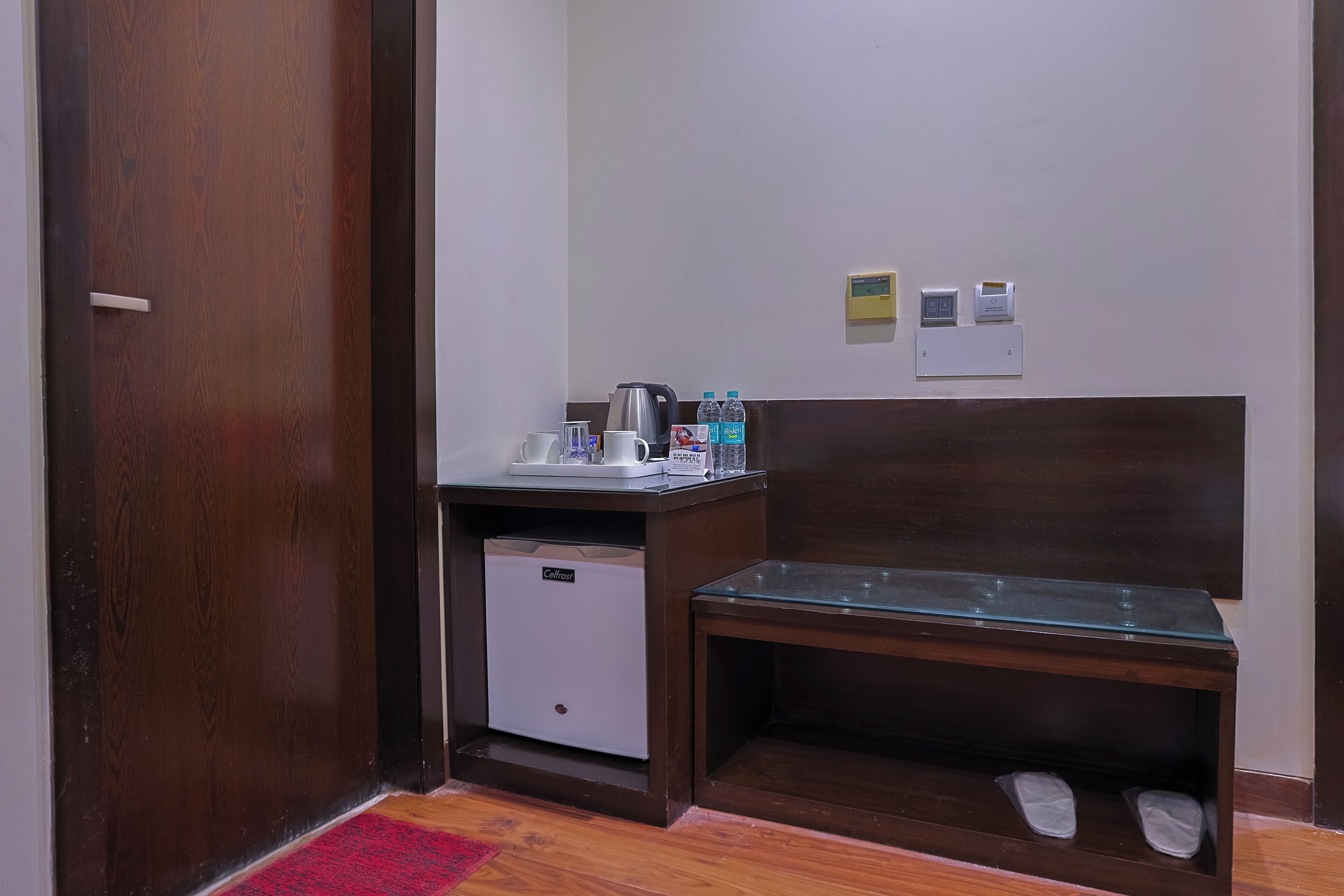 A hotel room with a wooden door, a small desk with a kettle, a glass table, a shoe rack, and a red rug.