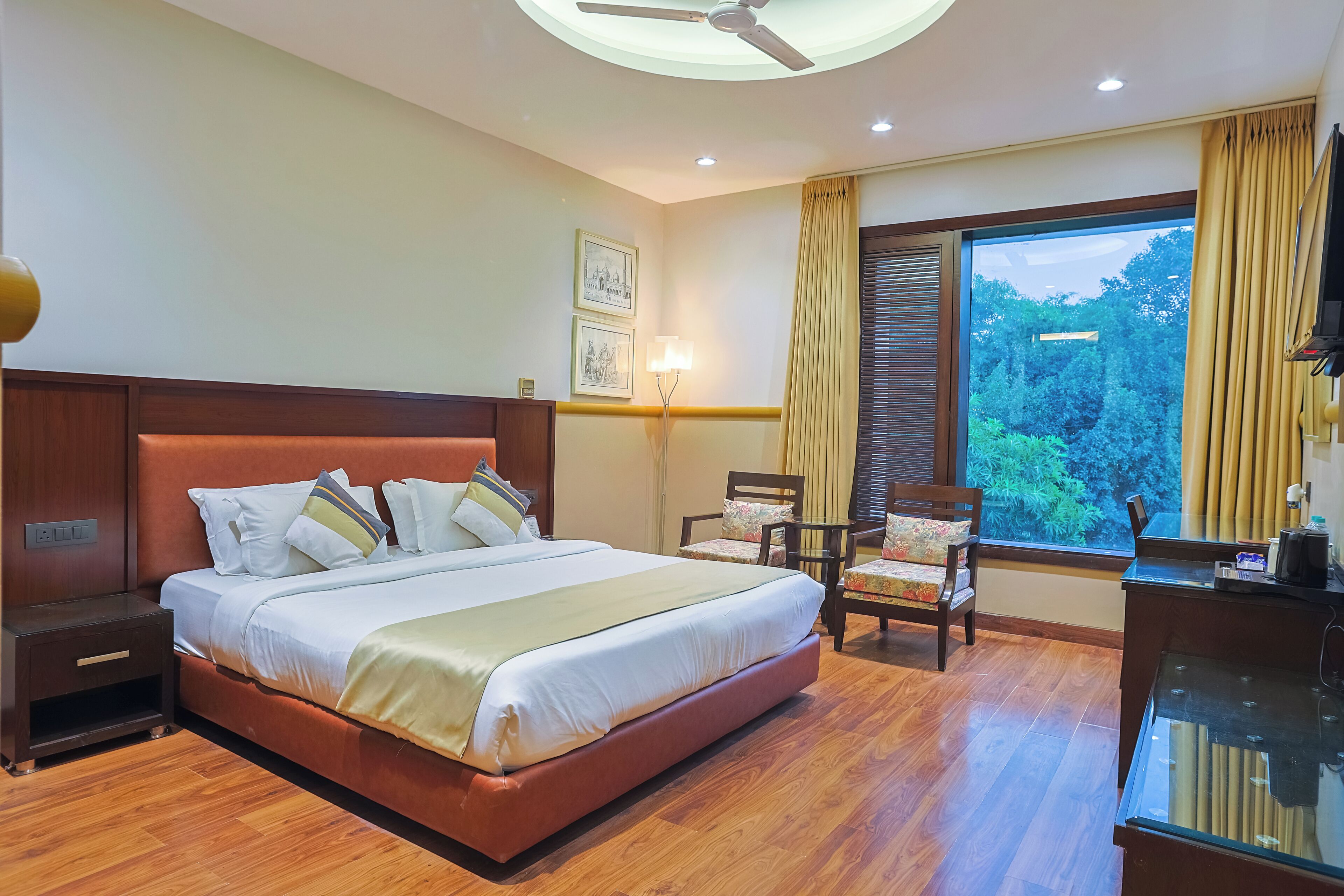 Quarto de hotel com uma cama grande, uma escrivaninha, uma cadeira, uma mesinha de cabeceira e uma janela com cortinas.