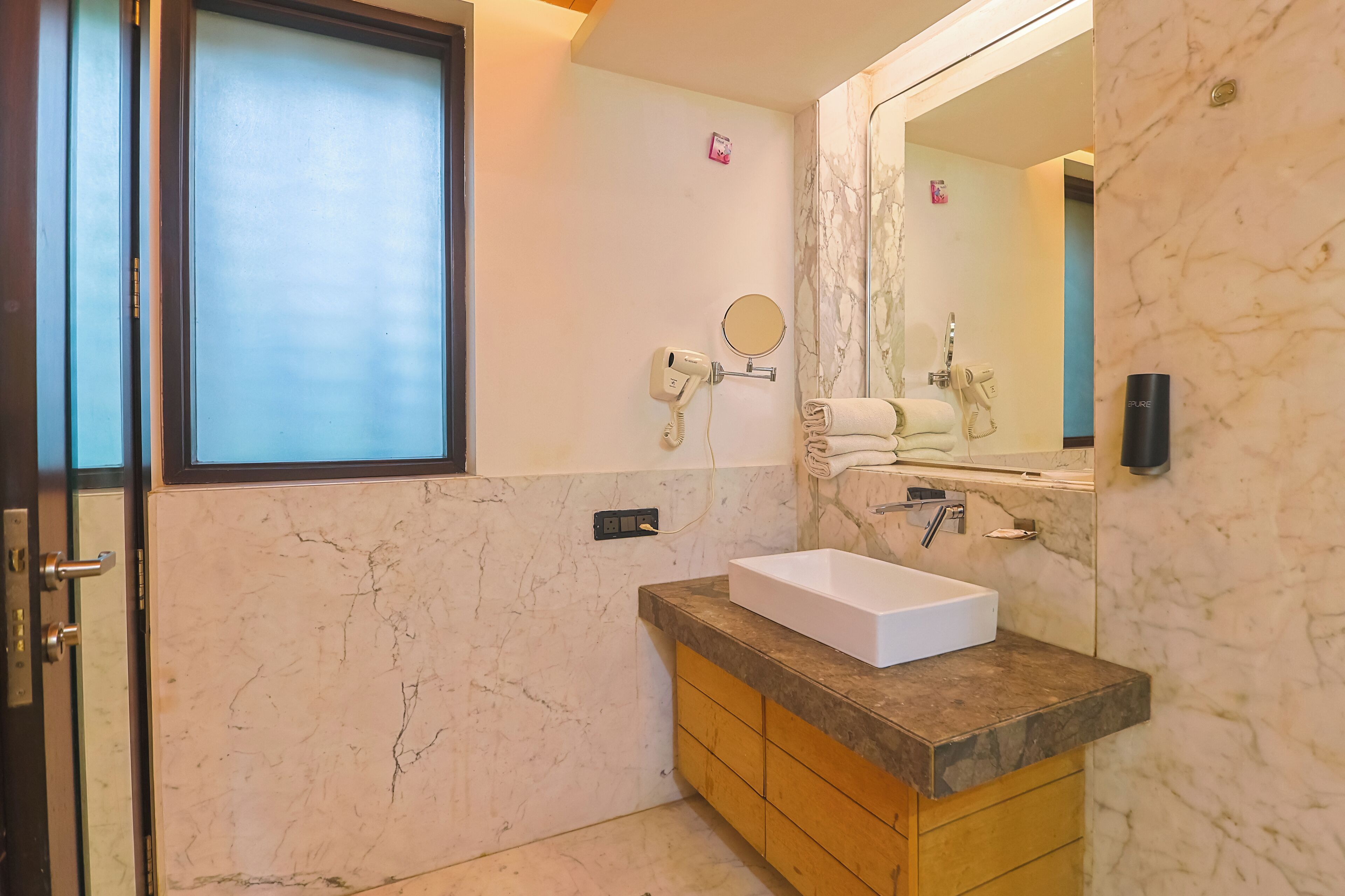 Un baño moderno con pared de mármol, un espejo grande, un lavamanos blanco y un perchero para toallas.
