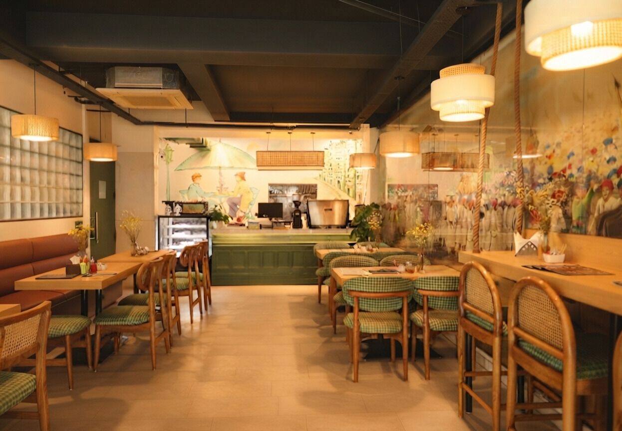 L’intérieur d’un restaurant avec des tables et des chaises en bois, une cuisine en arrière-plan et des luminaires suspendus.