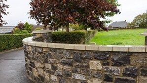 A stone wall with a curved top, a small tree with red leaves, and a grassy area with a house in the background. - Peaceful Rural Retreat with Mountain Views - Benbradagh Country Cottage (Dungiven)