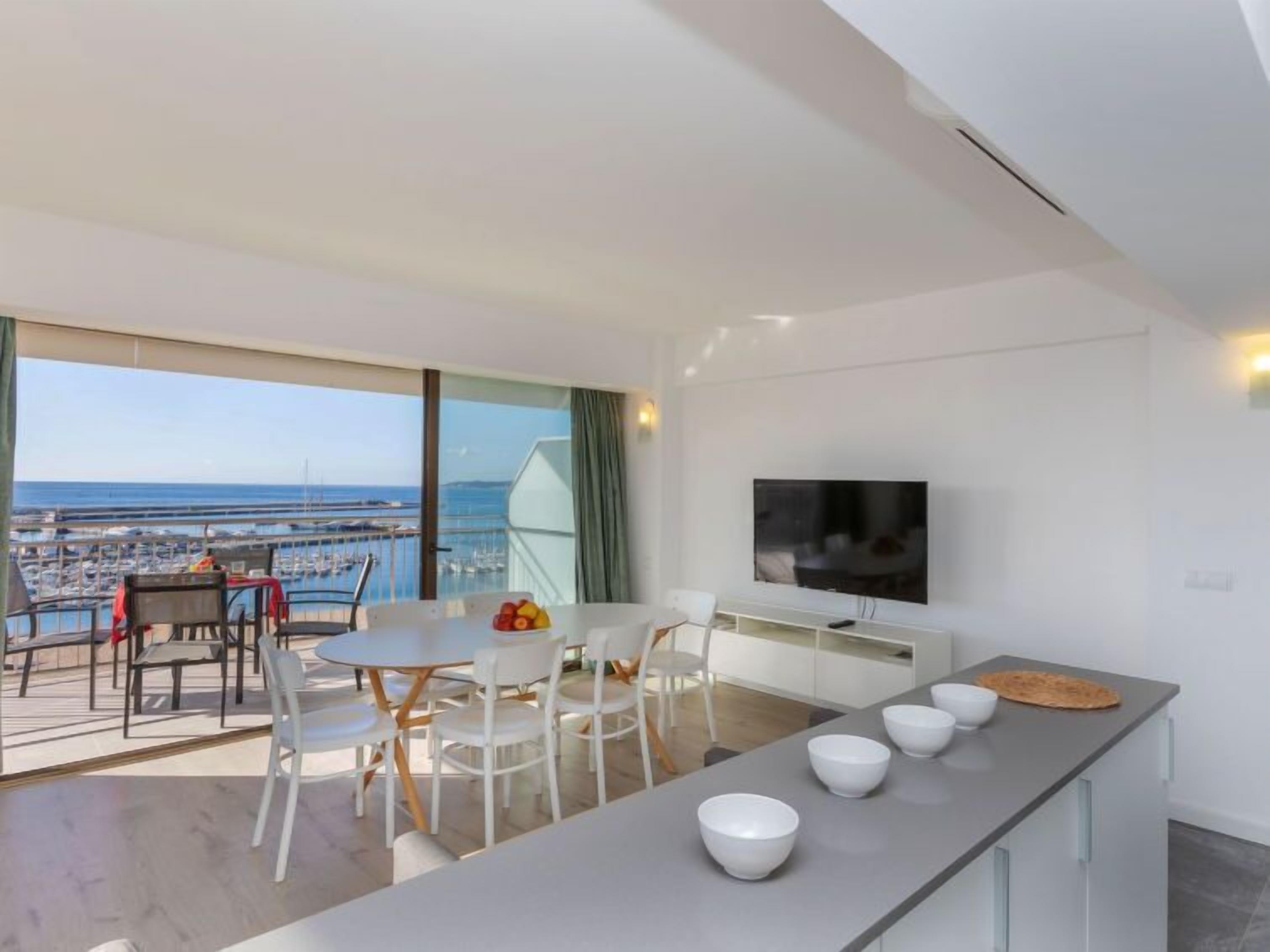 Un salon moderne doté d’un grand téléviseur, d’un coin repas avec une table et des chaises, et d’un balcon offrant une vue sur le port de plaisance.