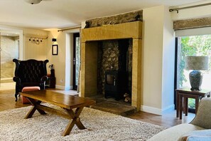 Cozy living room with a central fireplace, dark leather armchair, and wooden bench, offering a view of the surrounding greenery.