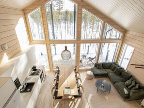 A spacious loft with a high ceiling, large windows offering a view of a forest, a dining area with a wooden table and chairs, and a cozy seating area with a sofa.