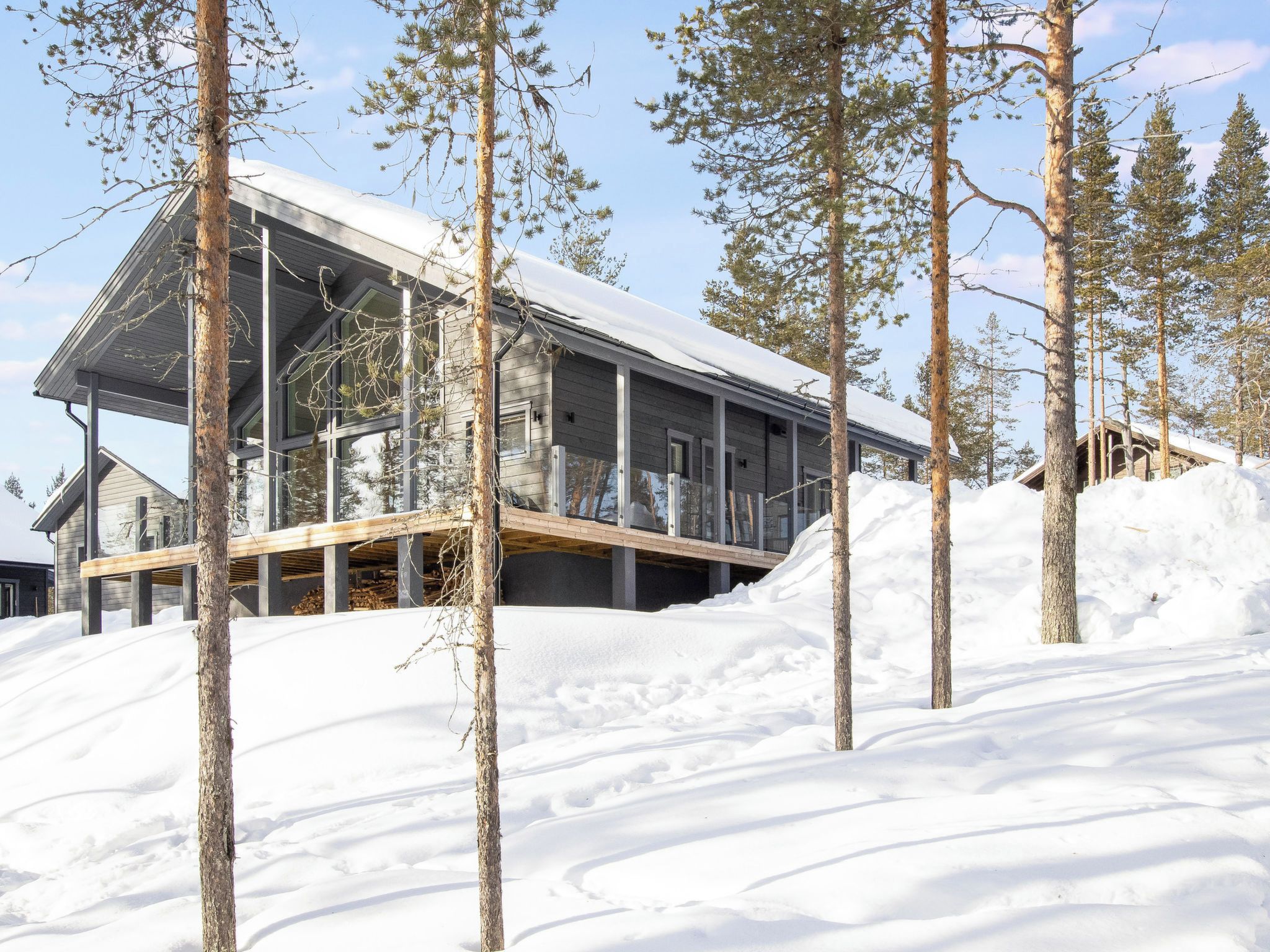 A modern cabin with a glass facade, surrounded by snow-covered ground and pine trees.