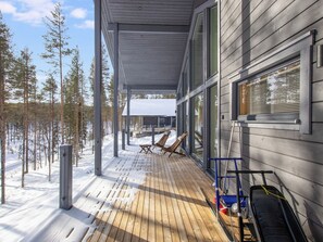 A covered wooden deck with two lounge chairs, a snow-covered ground, and a view of a forest.