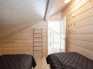 A small, cozy room with wooden walls and a sloped ceiling. There are two beds with dark bedding, a ladder against the wall, and a door leading to another room. - Villa velho by Interhome (Äkäslompolo)