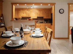 Un coin repas avec une table en bois dressée pour le repas, entourée d’armoires en bois et une cuisine en arrière-plan.
