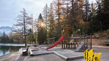 Ein Spielplatz mit einer roten Rutsche und Klettergerüsten, umgeben von Bäumen und einem See, mit Bergen im Hintergrund.