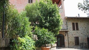 Une cour avec un grand arbre en pot, un mur de pierre et un bâtiment doté d’une façade en pierre et de volets en bois.