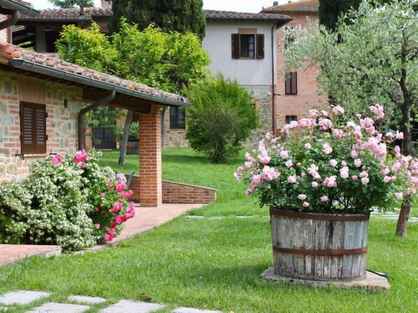 Un bâtiment en pierre avec un toit en tuiles, un jardinière en forme de tonneau en bois ornée de fleurs roses et une pelouse bien entretenue.
