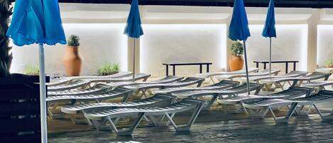 Un área junto a la piscina con sillas de lounge blancas y sombrillas azules, plantas en macetas y suelo de baldosas.