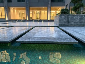 Un área de piscina moderna con superficie de agua cristalina, escalones de piedra que conducen a la entrada de un edificio y suelo de baldosas.