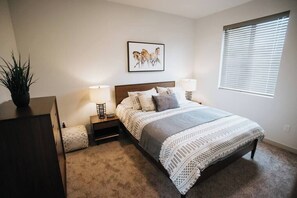 A bedroom with a bed, bedside tables, lamps, a wall-mounted picture, and a window with blinds.