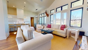 A spacious living room with a large window, a mounted deer head, and a kitchen in the background. - River City Resorts: Oxbow House, on the Guadalupe River (New Braunfels)