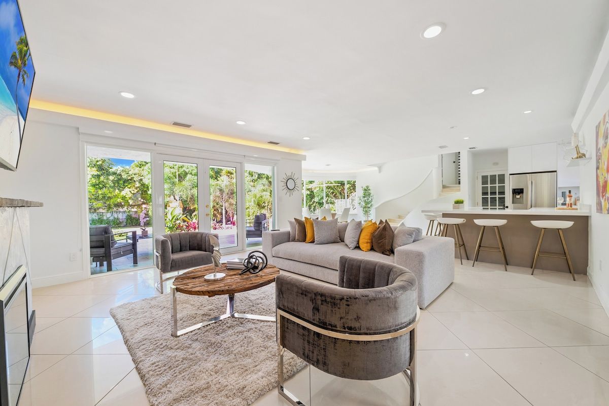A modern living room with a sofa, armchairs, and a coffee table. There is a kitchen area with a bar and stools.