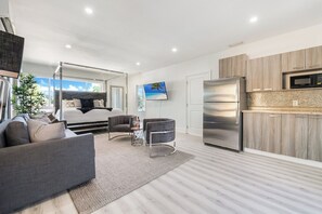 Ein moderner Wohnraum mit Sofa, Couchtisch, Fernseher, einem Bett mit Baldachin sowie einer Küchenzeile inklusive Kühlschrank und Mikrowelle.