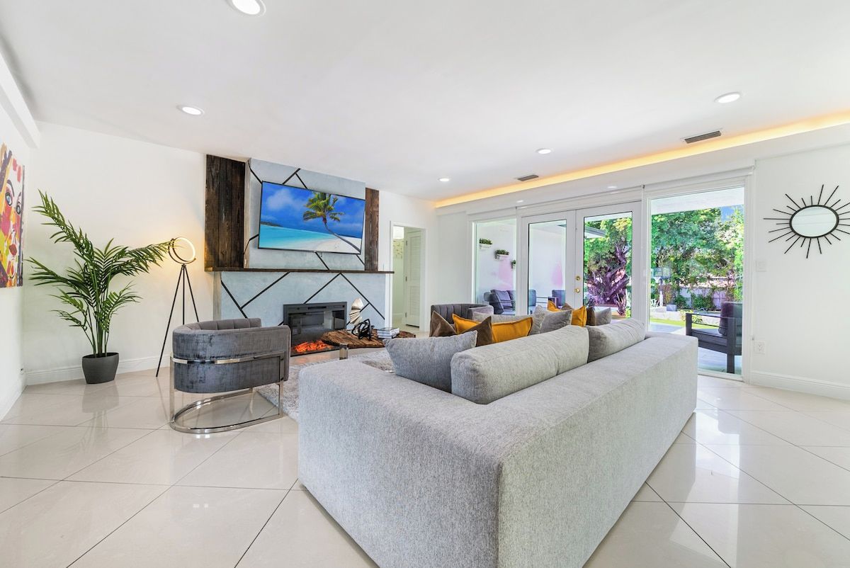 A modern living room with a large grey sofa, a fireplace, and a large window with a view of greenery.