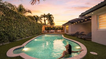 Una piscina con una persona nadando, rodeada de follaje y un edificio con terraza.