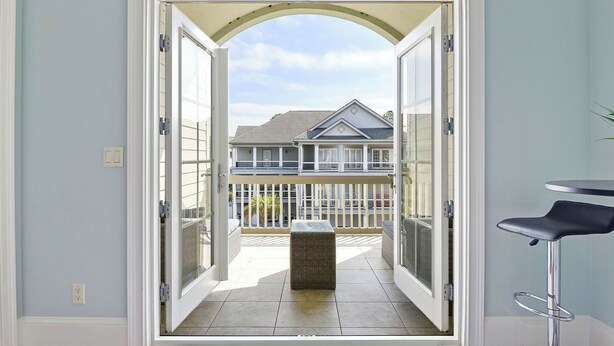 A view through a glass door into a balcony area with a wicker stool and a metal bar stool. - 5 Sandy Beach Trail- Beach House w/ Private Pool (Hilton Head)