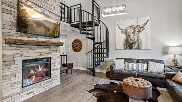 A modern living room with a stone fireplace, a black spiral staircase, a large cow painting, and a leather sofa.
