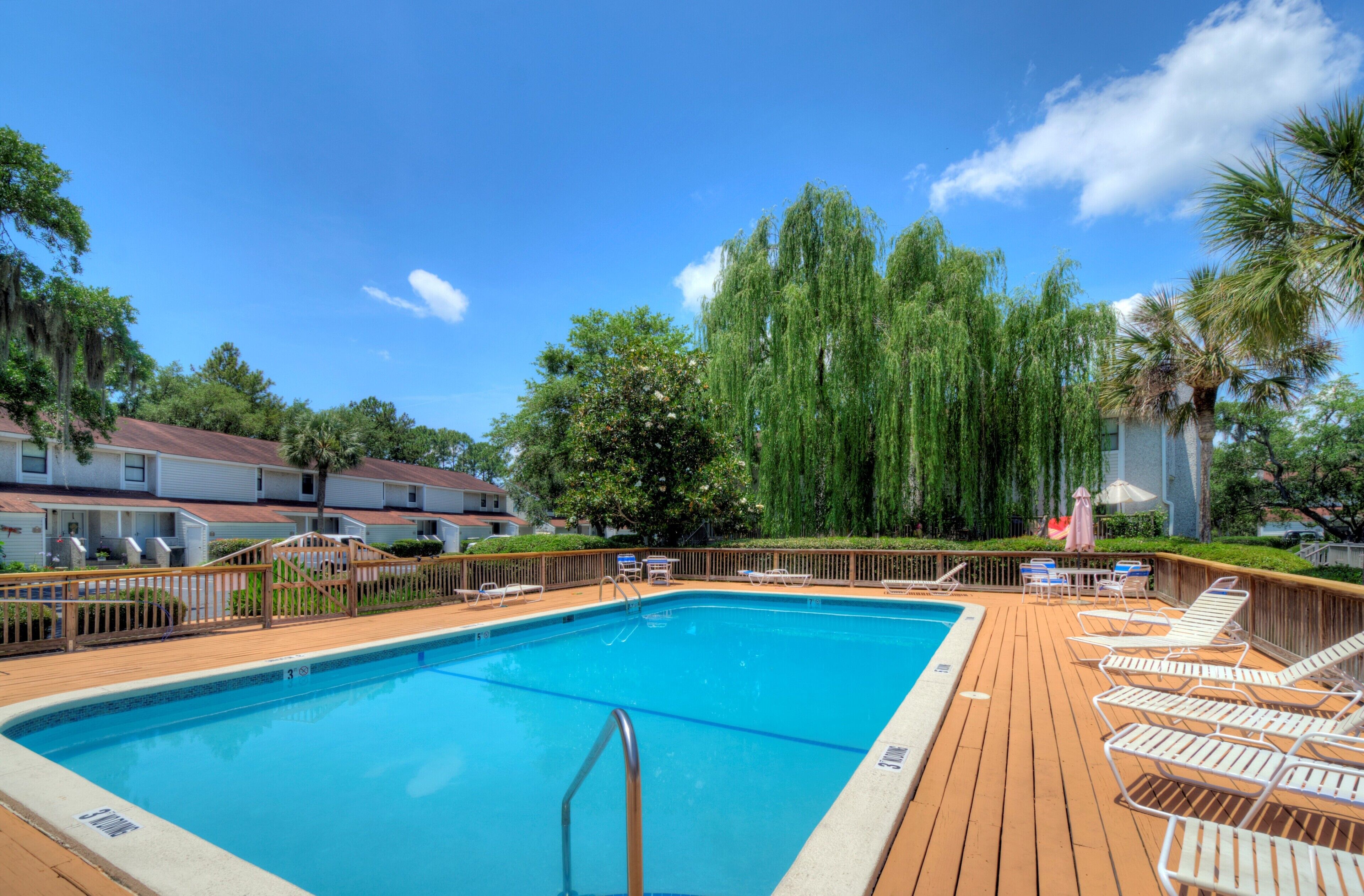A swimming pool with a wooden deck and lounge chairs, surrounded by trees and a building.