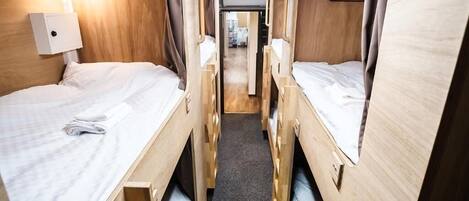 A narrow, well-lit corridor with bunk beds on either side, a central walkway, and a hanging coat rack.