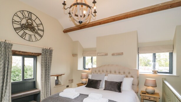 A bedroom with a large bed, a wooden ceiling beam, a wall clock, and a window with curtains. - The Dovecote (Denbigh)