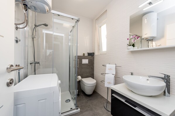 Une salle de bain moderne avec une douche à parois de verre, des toilettes blanches et un grand lavabo surmonté d’un miroir.