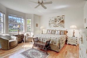 A bedroom with a bed, two nightstands, a desk, two chairs, a ceiling fan, and large windows.