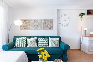 Un canapé de couleur turquoise agrémenté de coussins à pois et à motifs de papillons, une horloge blanche accrochée au mur et un vase rempli de fleurs jaunes posé sur le sol.