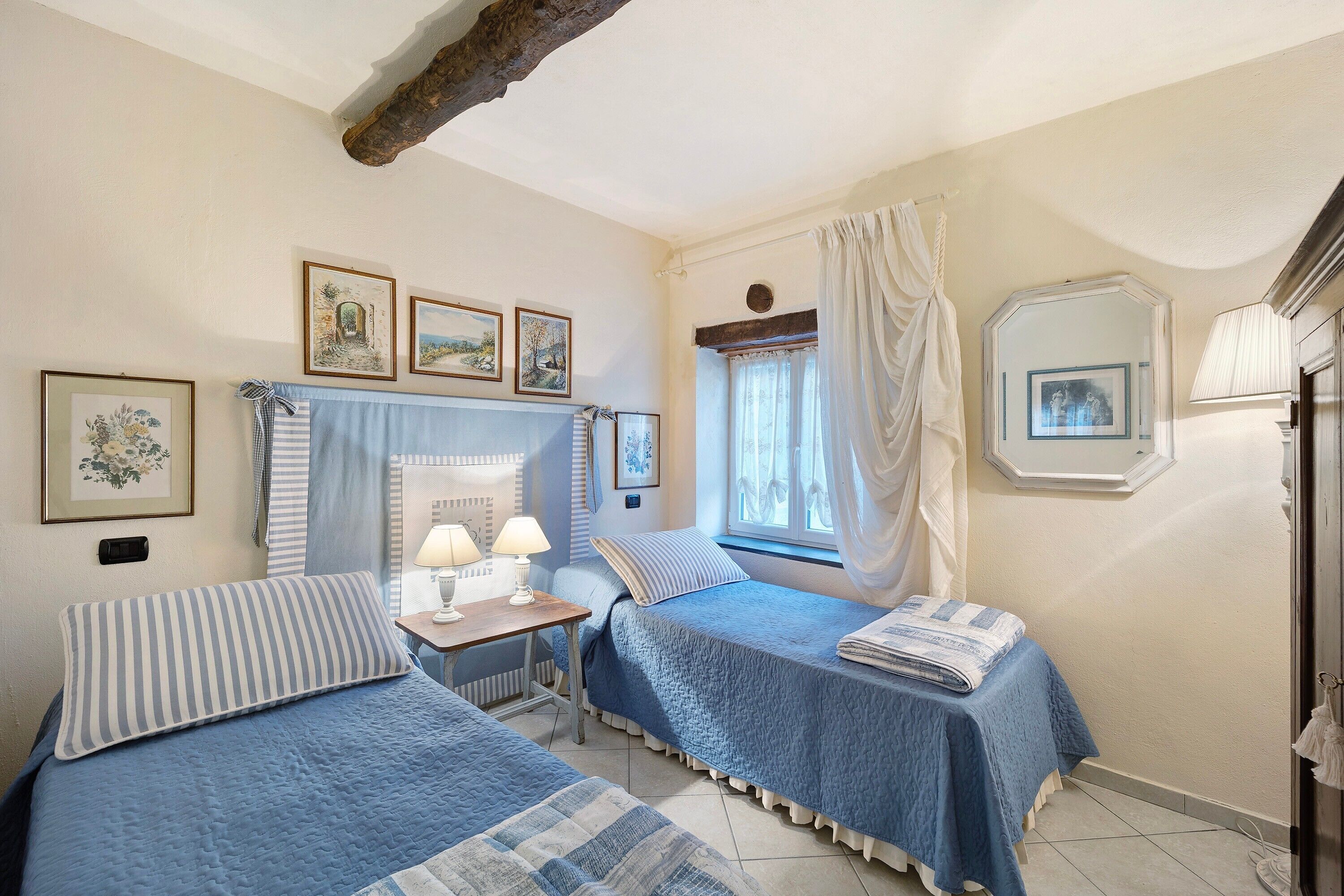 Une chambre avec deux lits, une table de chevet avec des lampes, une fenêtre avec des rideaux et des tableaux encadrés accrochés au mur.