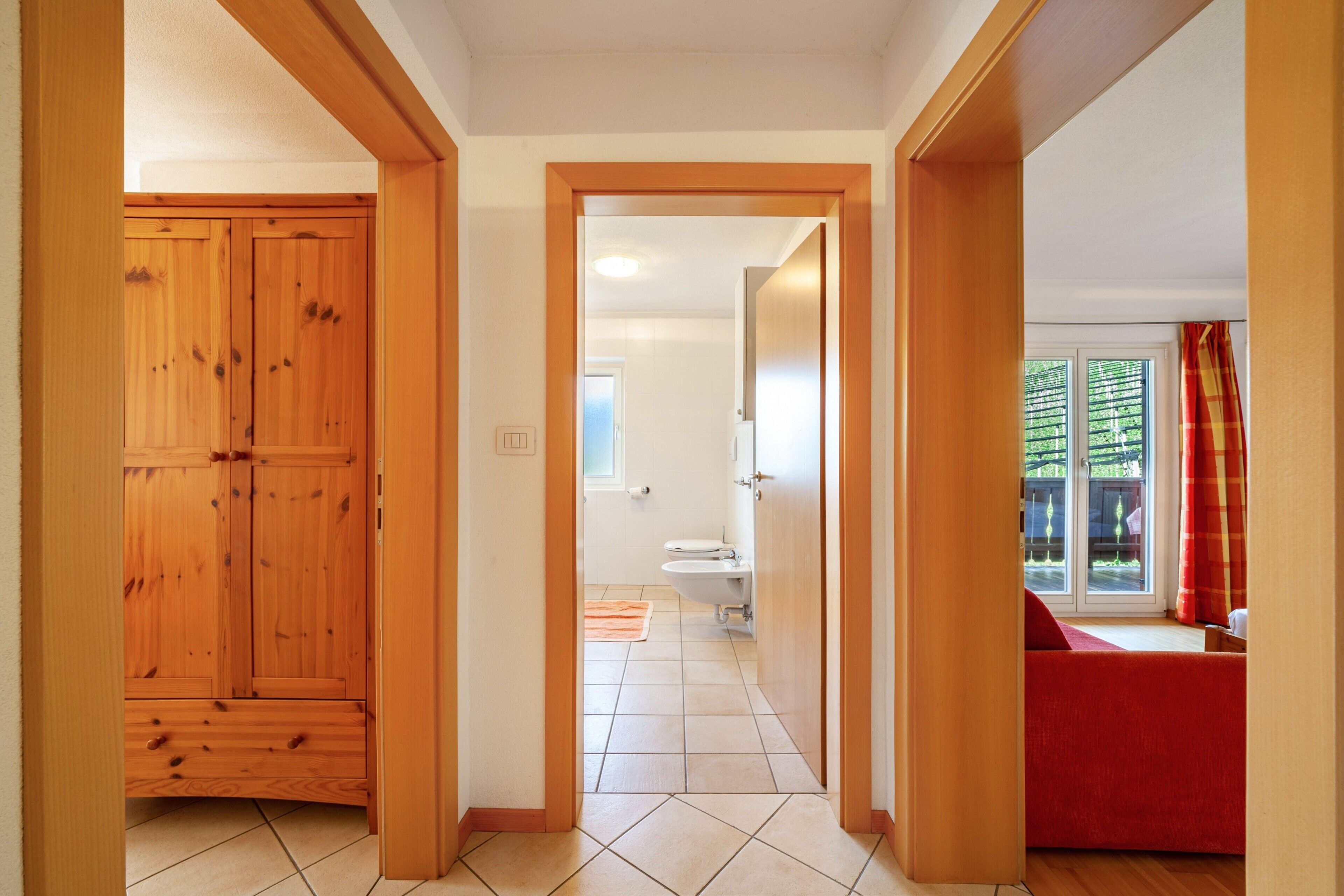 A hallway leading to a bathroom with a toilet and sink, wooden doors, and a red sofa in the adjacent room.