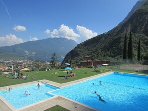 A swimming pool with people swimming and playing, surrounded by a grassy area with umbrellas and a view of a town and mountains in the background. - Apartment "Sara" with Lake View, Shared Pool & Wi-Fi (Riva del Garda)