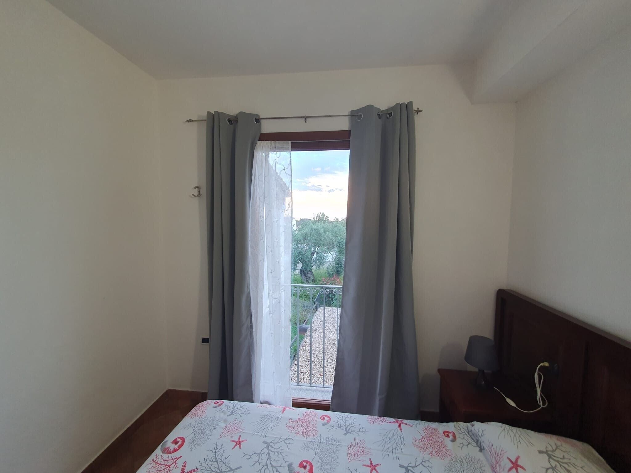 Une chambre à coucher avec un lit, une table de chevet, une fenêtre avec des rideaux et une vue sur un balcon et des arbres.